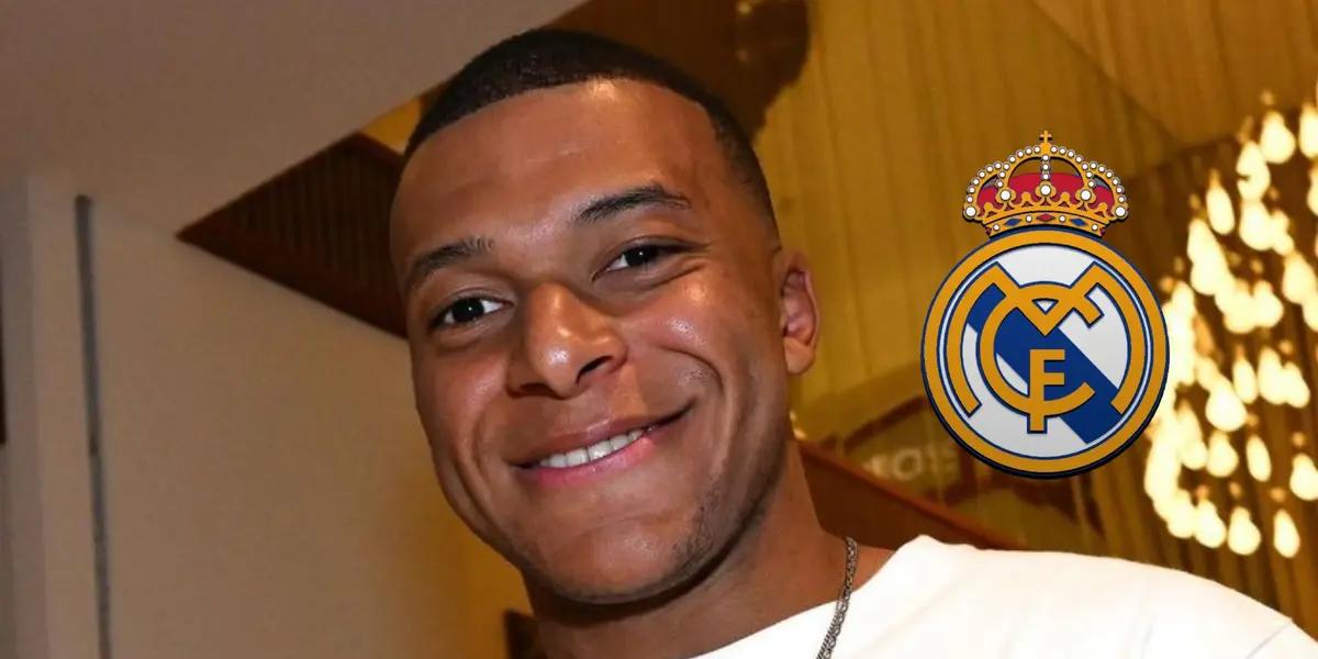Kylian Mbappé smiles wearing normal clothes while the Real Madrid badge is next to him.