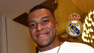 Kylian Mbappé smiles wearing normal clothes while the Real Madrid badge is next to him.